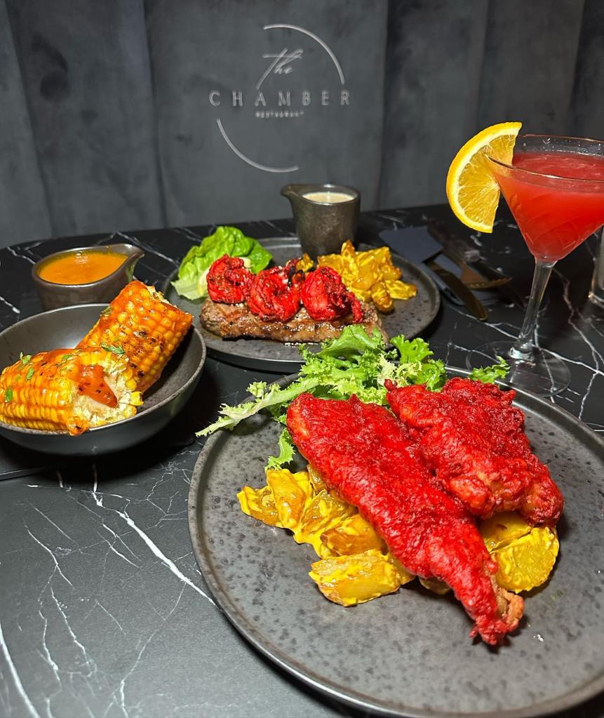 a fusion of flavours with an indian twist. masala fish and chips, the fish is a red colour batter and creamy chips. in a separate bowl is charred corn on the cob, and a separate helping of suft and turf.