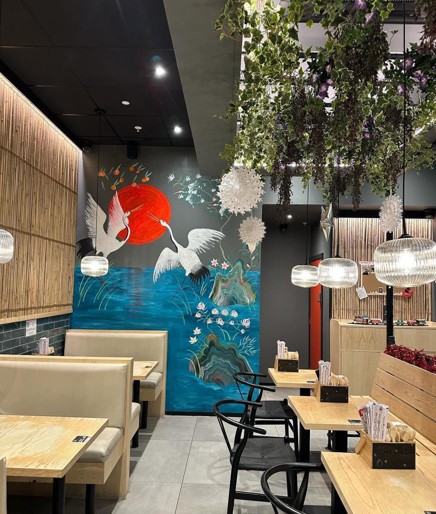 The Grounded Kitchen in Metrocentre boasts an inviting interior, featuring a hand-painted mural of storks flying over water with a sunset backdrop. Green plants dangle from the ceiling, and textured light fixtures contribute to a serene and cozy atmosphere.
