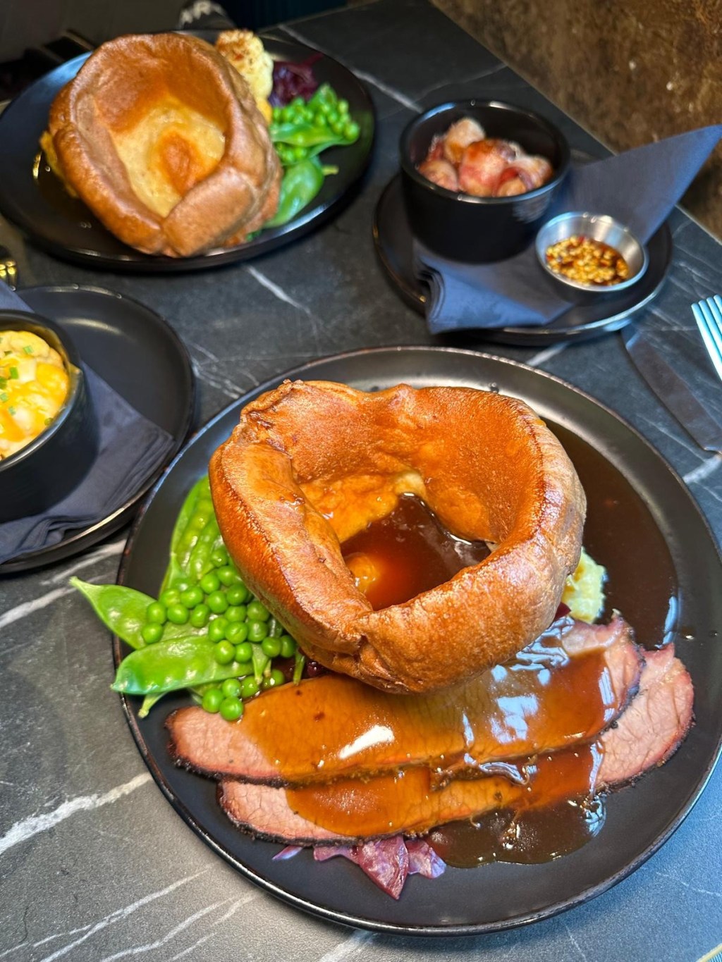 Tender beef brisket served with seasonal vegetables and crispy roast potatoes, also cheesy cauliflower, succulent pigs in blankets and sweet chili dip all laid out on a black marble table