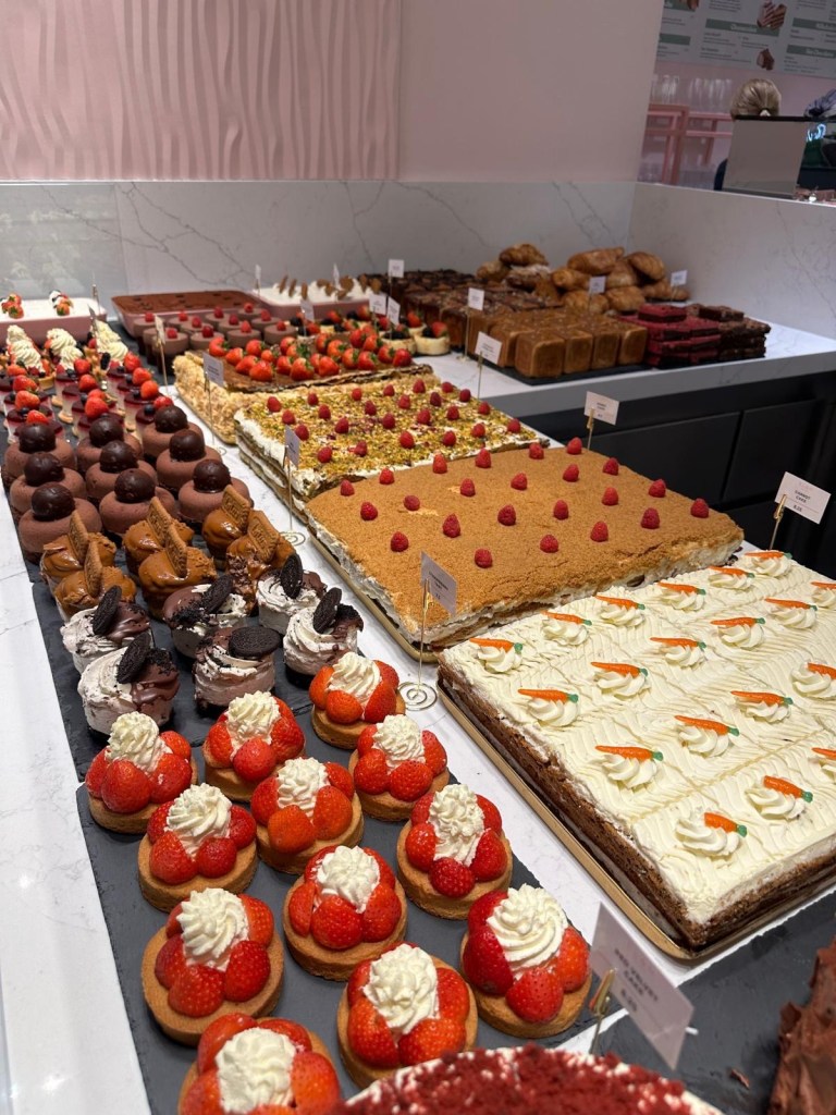 A stylish café space with light colors, modern lighting, neon signs, and lush greenery and a selection of world class cakes on display