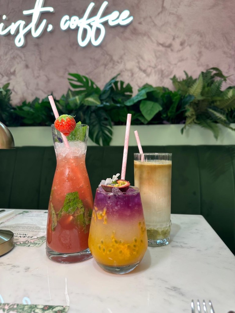 Ombré Passion Fruit Cooler: A refreshing drink layered with golden passion fruit and ombré hues, served in a tall glass with ice. Strawberry Chemex Mojito: A vibrant pink drink with fresh strawberries, mint leaves, and a light fizz, served in a chemex glass. Pistachio Spanish Latte: A creamy latte with a hint of green pistachio, topped with frothy almond milk foam, served in a clear glass.