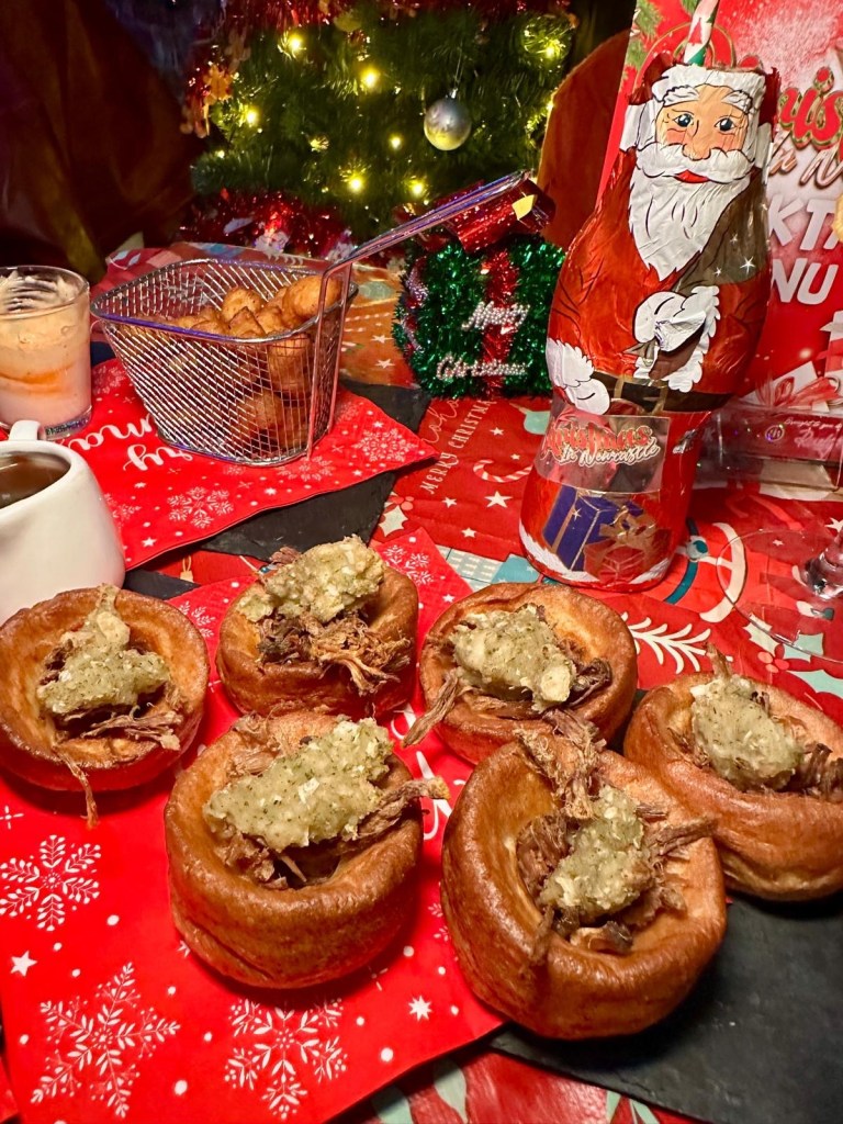 Three festive nibbles: crispy Mini Yorkshire Puddings filled with slow-roasted beef, stuffing, and gravy, served on a rustic plate. Golden Baked Camembert Rounds, crispy on the outside and gooey inside, paired with a festive garnish. A plate of crispy Cauliflower Wings drizzled with vegan sriracha mayonnaise, served as a vibrant festive snack.