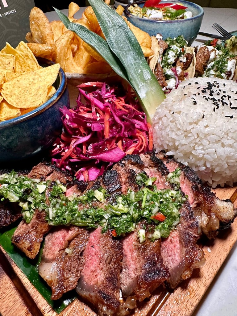 Steak Sharing Board: Tender birria strip loin steaks with steak tacos, chimichurri, pickled slaw, sticky rice, corn luu, and crispy potato sidewinders.