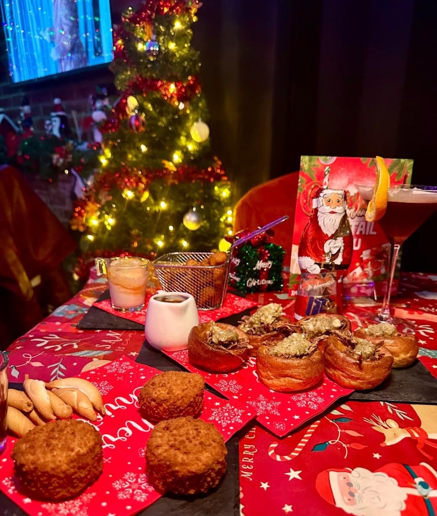 Three festive nibbles: crispy Mini Yorkshire Puddings filled with slow-roasted beef, stuffing, and gravy, served on a rustic plate. Golden Baked Camembert Rounds, crispy on the outside and gooey inside, paired with a festive garnish. A plate of crispy Cauliflower Wings drizzled with vegan sriracha mayonnaise, served as a vibrant festive snack.