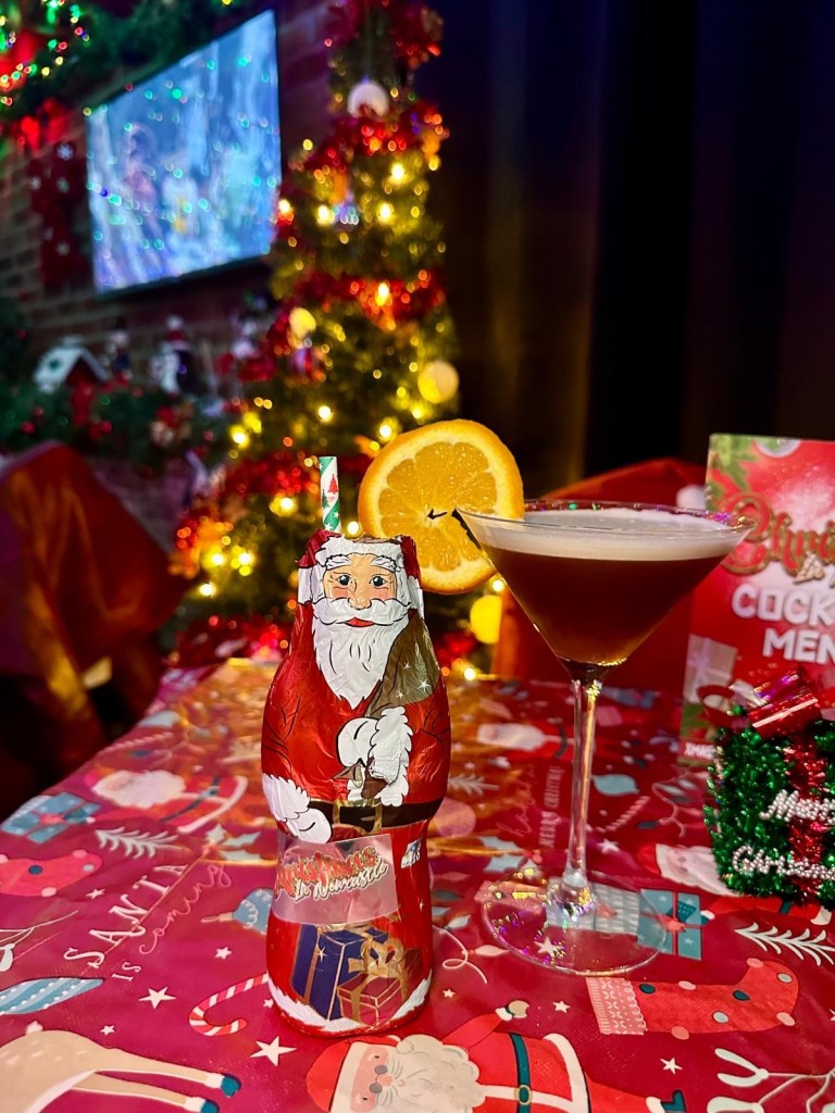 A Gingerbread Martini served in a hollow chocolate Santa, decorated with festive flair, ready to enjoy and a Spiced Carajillo cocktail featuring tequila with a festive twist, garnished with a citrus peel. A Gingerbread Martini served in a hollow chocolate Santa, decorated with festive flair, ready to enjoy