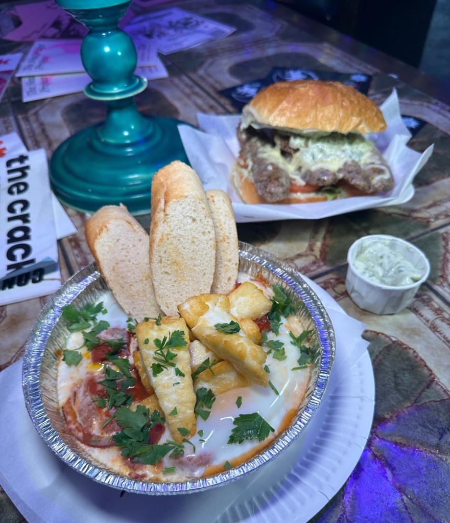 A mouthwatering display of Tunisian Eggs in a spicy tomato and chili sauce, served with crusty bread and halloumi on the side, alongside an Italian SLT sandwich filled with sweet Italian sausage, fresh spinach, and pesto aioli.
