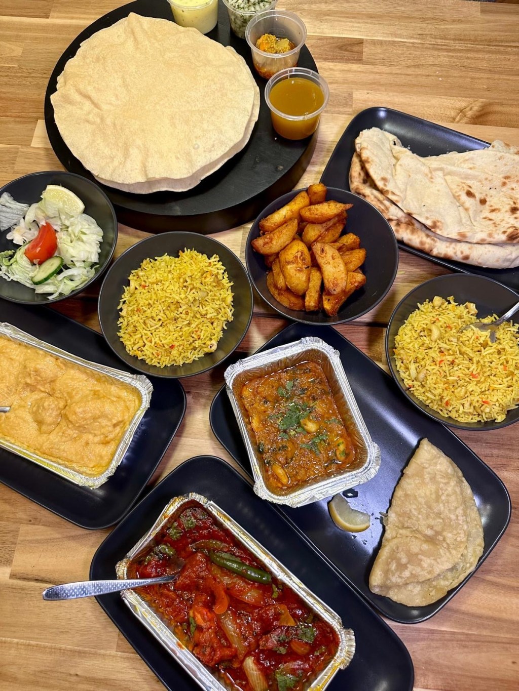 Aerial view of an Indian feast featuring popadoms with condiments, Bhuna Prawn Puree, King Prawn Jalfrezi, Chicken Butter Cream, Spicy Garlic Chips, Nut Pilau, and Peshwari Naan.