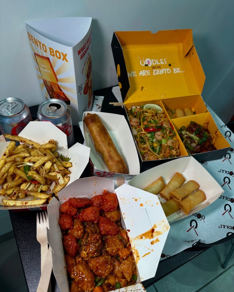 A variety of dishes from Oodles Wok, including a Bento Box with egg fried noodles, prawns, veggie spring rolls, Oodles signature wings, salt & pepper chips, Udon noodles with sweet & sour chicken and fiery garlic chicken, a jumbo roll, and fortune cookies.