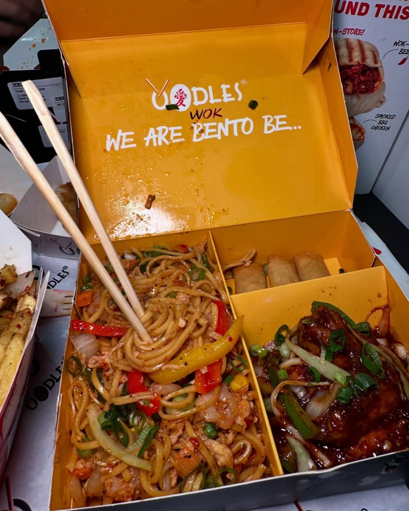 Bento box with egg fried noodles, stir-fried vegetables, prawns, veggie spring rolls, Oodles signature wings, and salt & pepper chips.