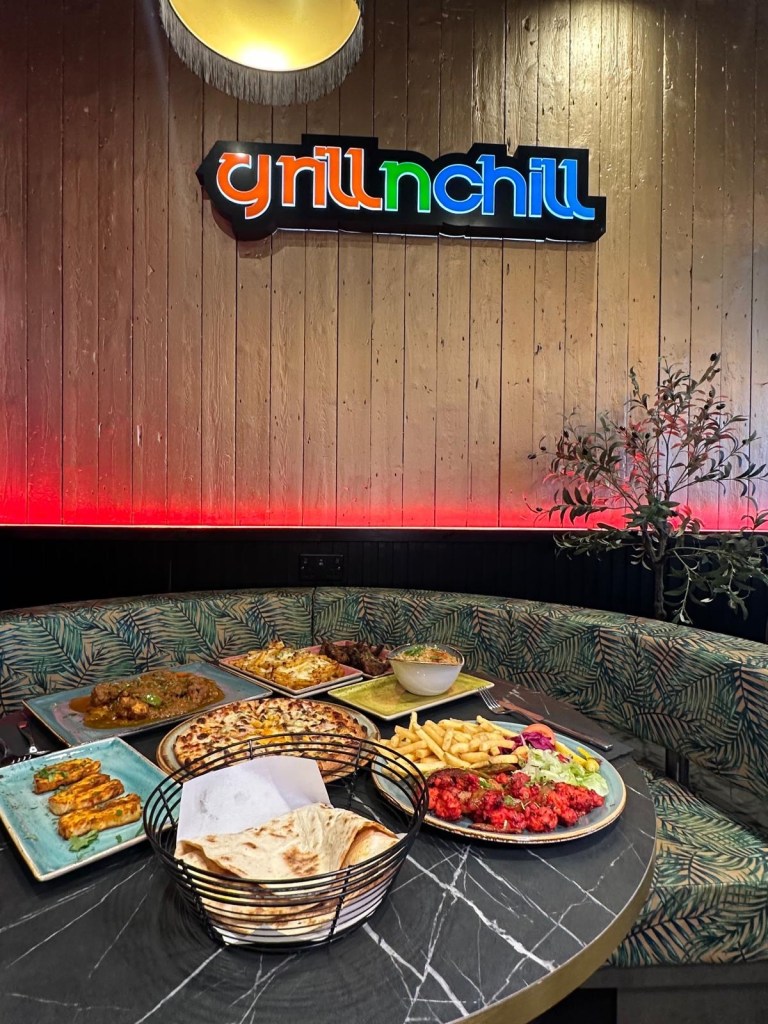 A booth table filled with a variety of dishes, including Chicken Boti Curry with naan, grilled lamb chops, a vegetarian pizza, halloumi slices, a king prawn kebab with side salad, and cheesy chips with melted cheese and vegetables