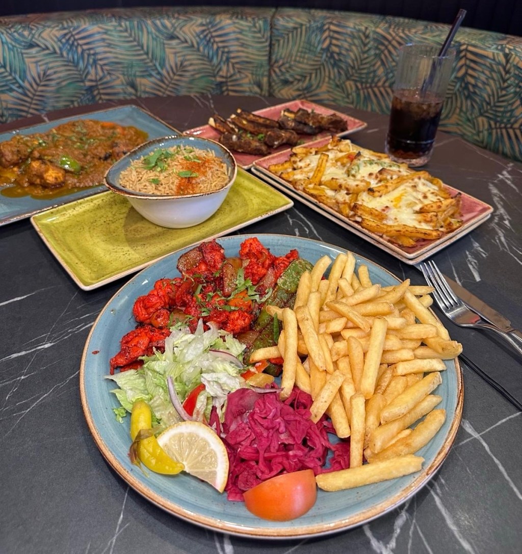 A booth table filled with a variety of dishes, including Chicken Boti Curry with naan, grilled lamb chops, a vegetarian pizza, halloumi slices, a king prawn kebab with side salad, and cheesy chips with melted cheese and vegetables