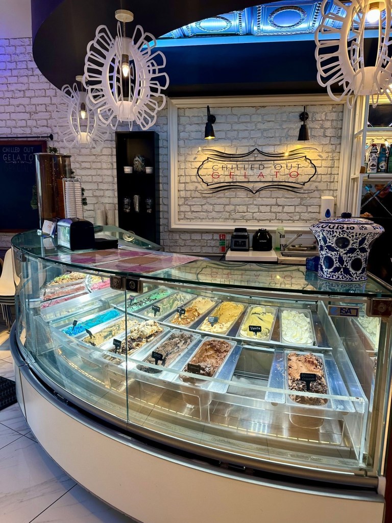 A display case showcasing a variety of colorful gelato flavors, with toppings and waffle baskets neatly arranged