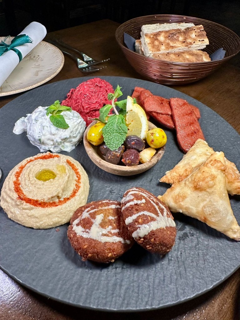 An elegant Turkish mezze platter with sucuk, falafel, muska boregi, humus, beetroot salad, cacik, olives, and freshly baked sesame seed bread, served on a polished wooden table