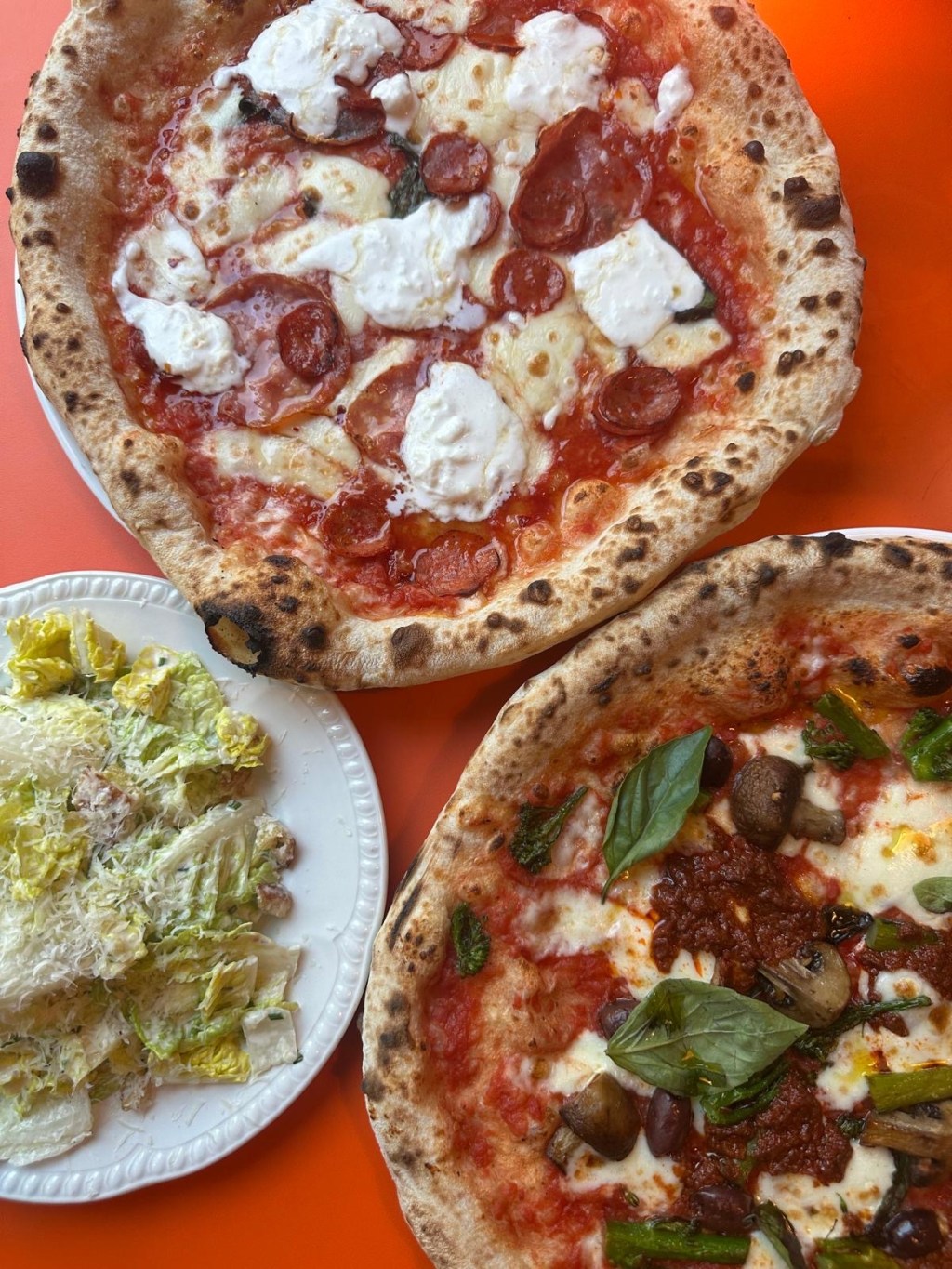A table filled with a variety of Rudy's dishes, including Caesar salad, 'Nduja Ortolana pizza, Carni pizza, tiramisu, and a Mandarino Spritz, alongside espresso and oat milk latte