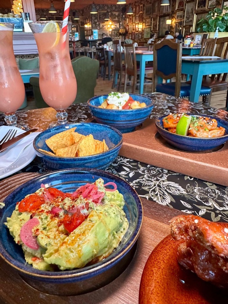 A bowl of smooth guacamole topped with coriander pesto, roasted peppers, and pickled red onion, served with crispy tortilla chips.