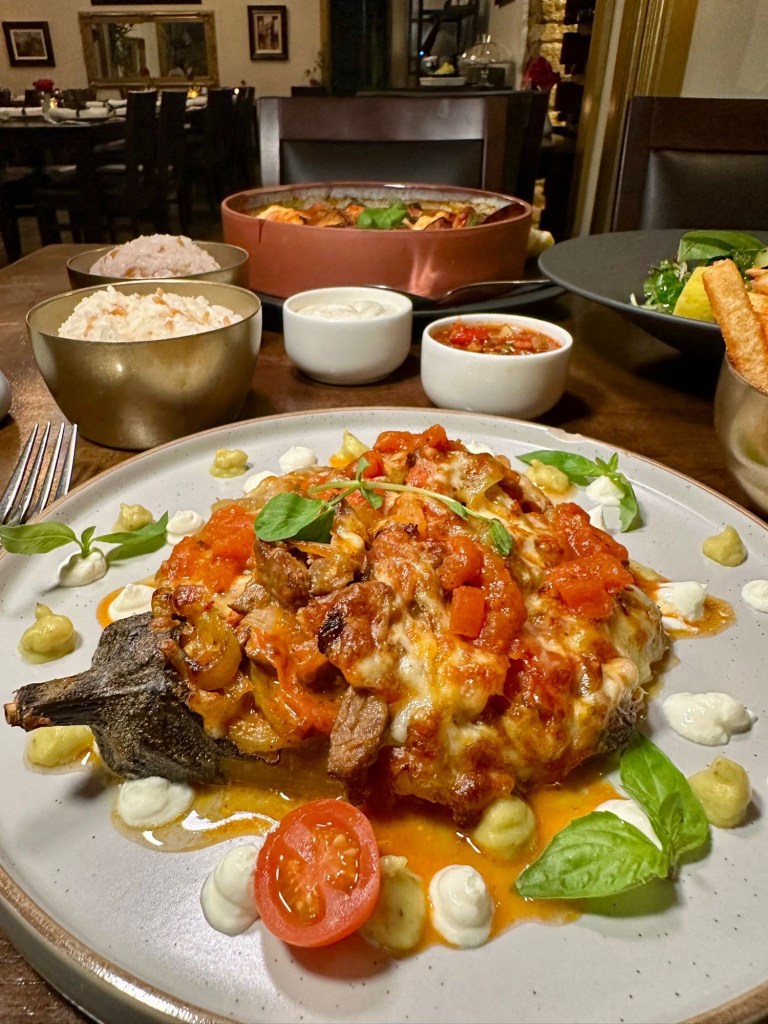 Grilled aubergine stuffed with juicy lamb, topped with creamy béchamel sauce and melted cheese, served with a garnish of fresh greens.