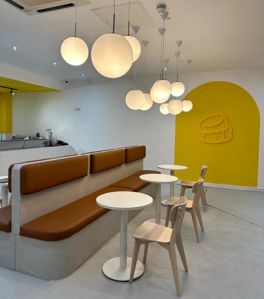 A modern and cozy café interior at Fluffy Fluffy, with soft lighting, minimalistic wooden furniture, and an open kitchen showcasing pancake preparation.