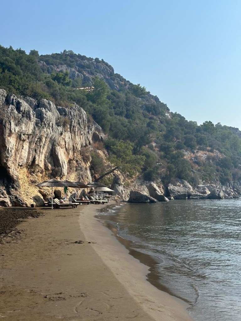A tranquil private beach at TUI Blue Seno, with golden sand, clear turquoise waters, and sun loungers shaded by parasols, framed by lush greenery and distant hills.