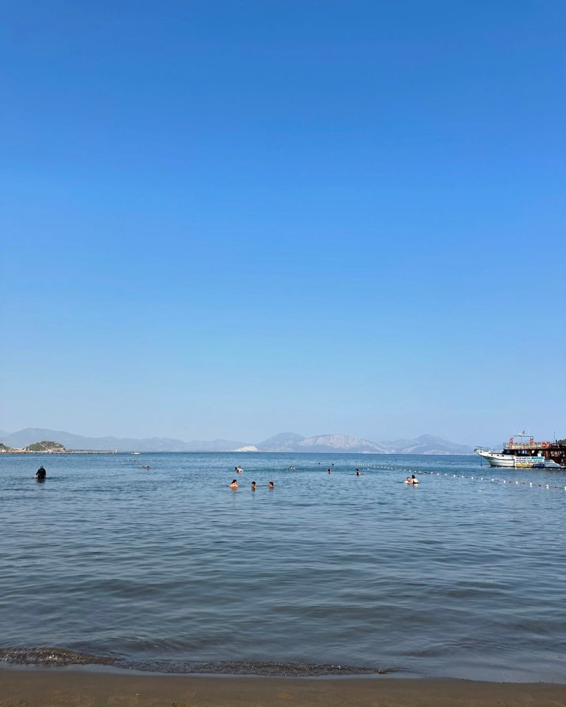 A tranquil private beach at TUI Blue Seno, with golden sand, clear turquoise waters, and sun loungers shaded by parasols, framed by lush greenery and distant hills.