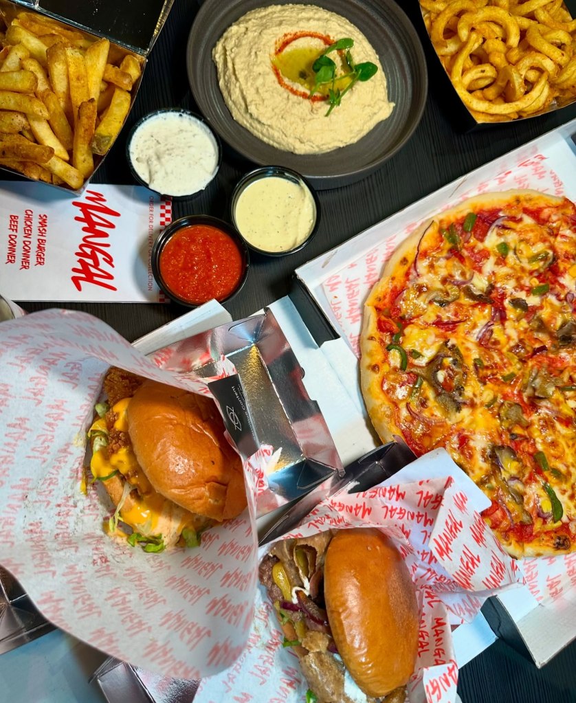 A feast of Mangal’s signature dishes, including burgers, pizza, fries, hummus, and chili sauce, all beautifully arranged on a table.
