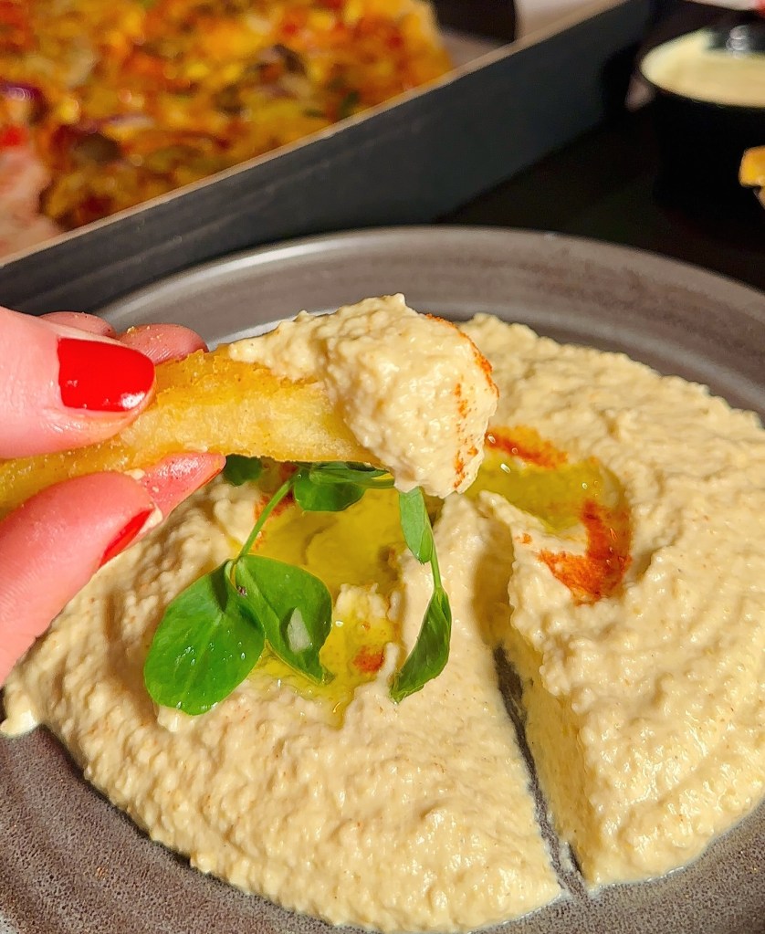 A creamy portion of hummus with a drizzle of olive oil, alongside a vibrant red chili sauce with a rich, spicy kick.