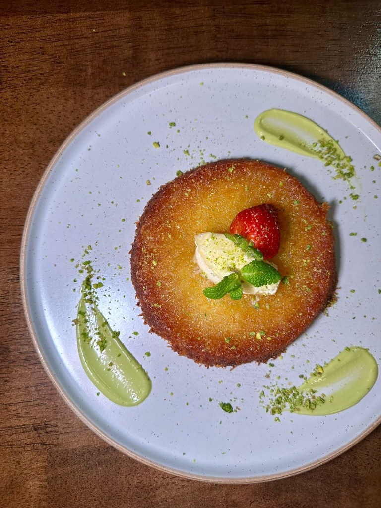 Golden kunefe dessert topped with a layer of pistachios and clotted cream, served on a plate with syrup drizzle