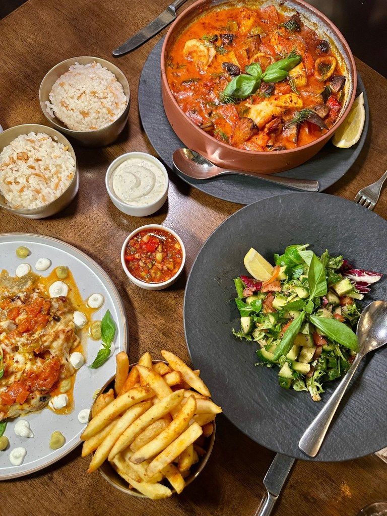 A vibrant spread of Turkish starters and mains, featuring a mezze platter with falafel, muska boregi, humus, beetroot salad, cacik, and freshly baked Turkish bread; alongside mains like Bostan Kebab with grilled aubergine stuffed with lamb and melted cheese, a seafood casserole with prawns, mussels, squid, and salmon in a creamy tomato-garlic sauce, and a fresh Mediterranean salad with pomegranate molasses dressing