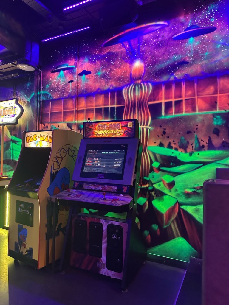 A row of classic arcade machines glowing under neon lights, featuring games like Tetris, Tekken, and Mortal Kombat at NQ64 Newcastle.