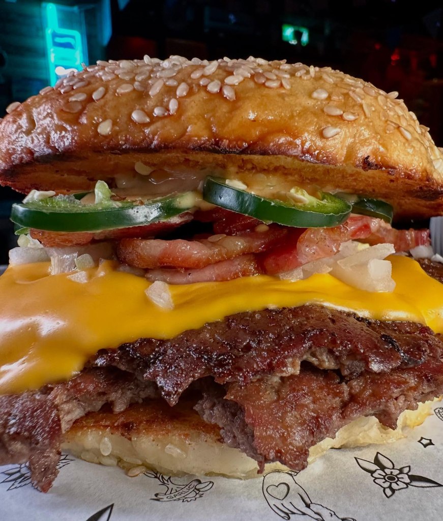 A double beef patty burger with American cheese, smoked bacon, fresh jalapenos, diced onions, and house sauce in a toasted seeded bun.