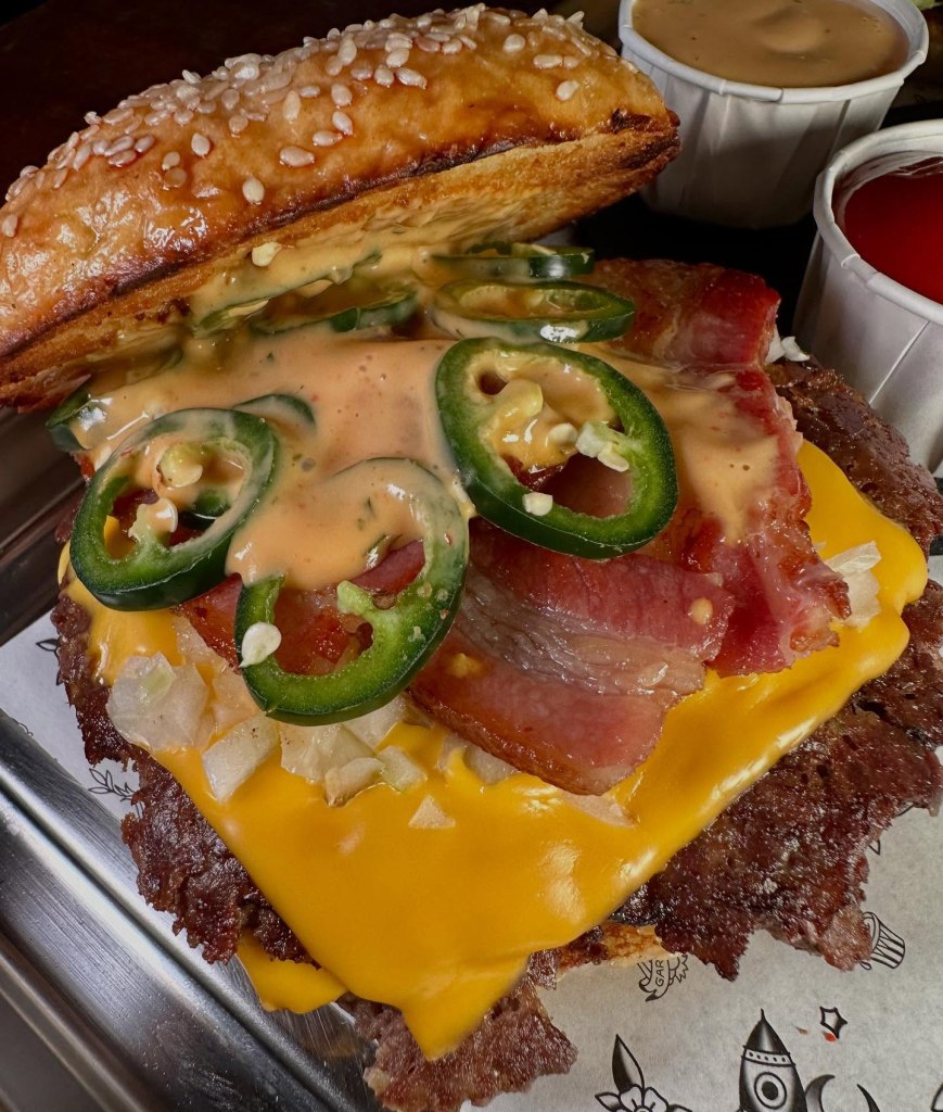 A double beef patty burger with American cheese, smoked bacon, fresh jalapenos, diced onions, and house sauce in a toasted seeded bun.