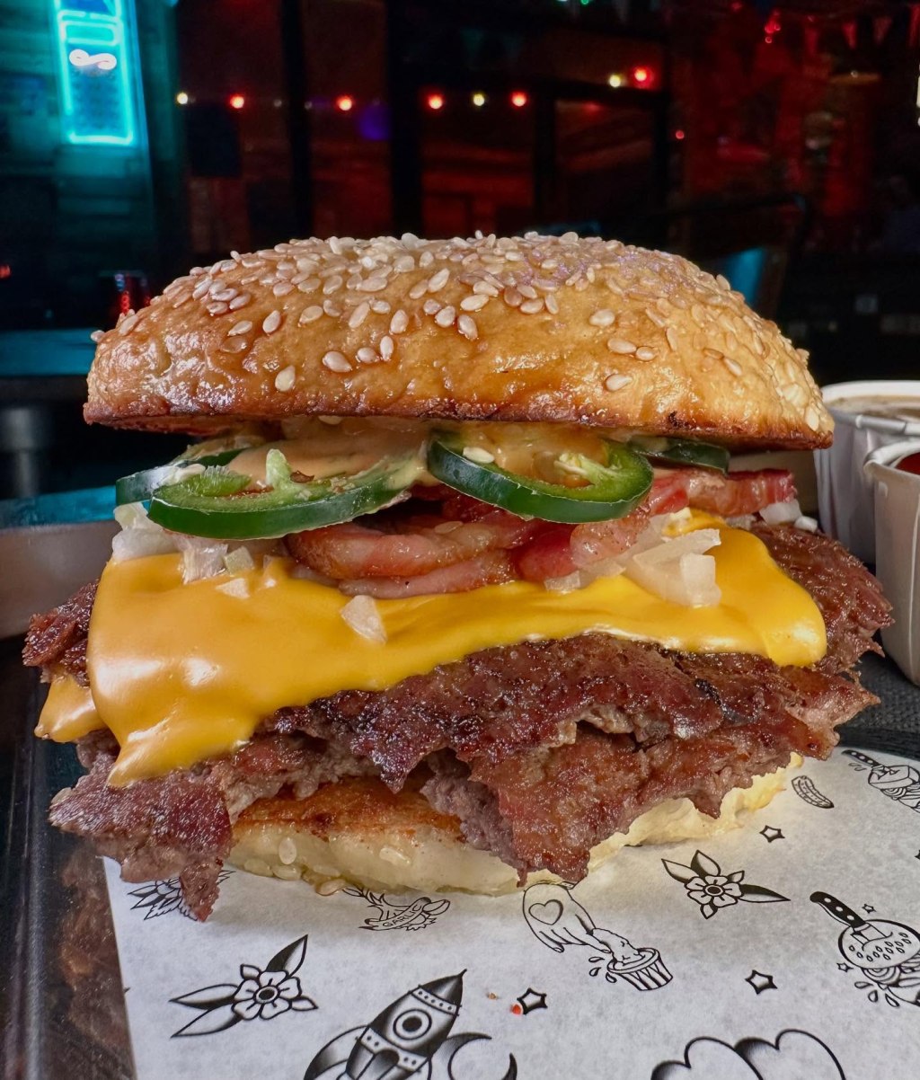 A double beef patty burger with American cheese, smoked bacon, fresh jalapenos, diced onions, and house sauce in a toasted seeded bun.