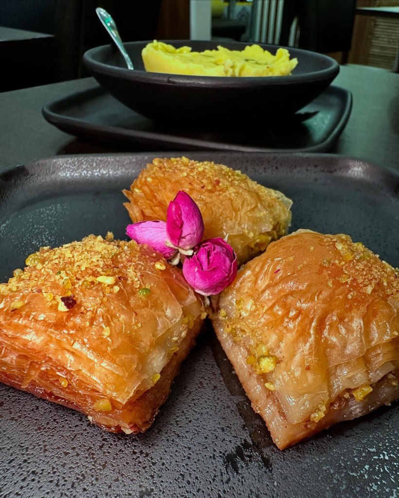 A portion of golden, flaky baklava topped with crushed pistachios and garnished with tiny roses.