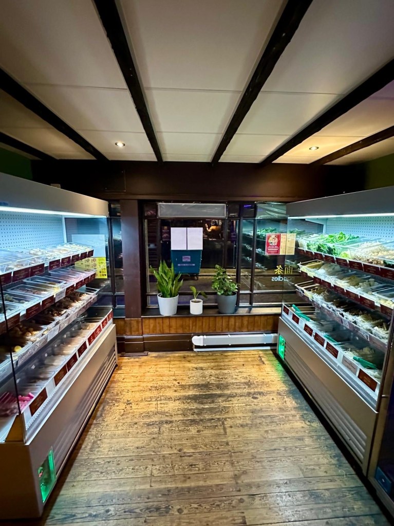 A row of refrigerated shelves stocked with fresh ingredients, including leafy greens, mushrooms, seafood, meats, and noodles, ready for selection.