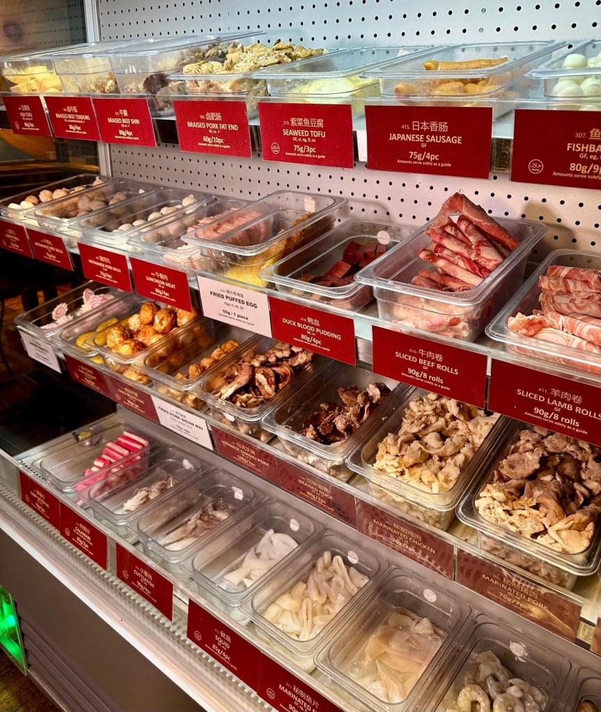 A row of refrigerated shelves stocked with fresh ingredients, including leafy greens, mushrooms, seafood, meats, and noodles, ready for selection.