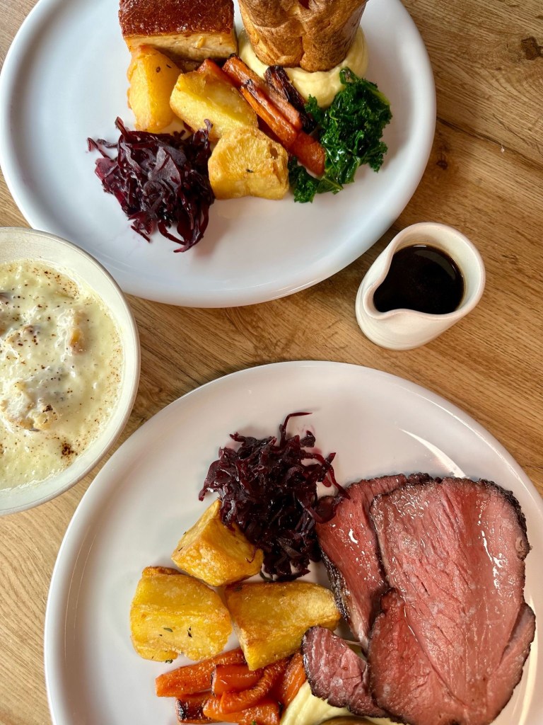 A hearty Sunday roast spread featuring roast beef, crispy pork belly, Yorkshire puddings, roast potatoes, seasonal vegetables, and a bubbling dish of cauliflower cheese.