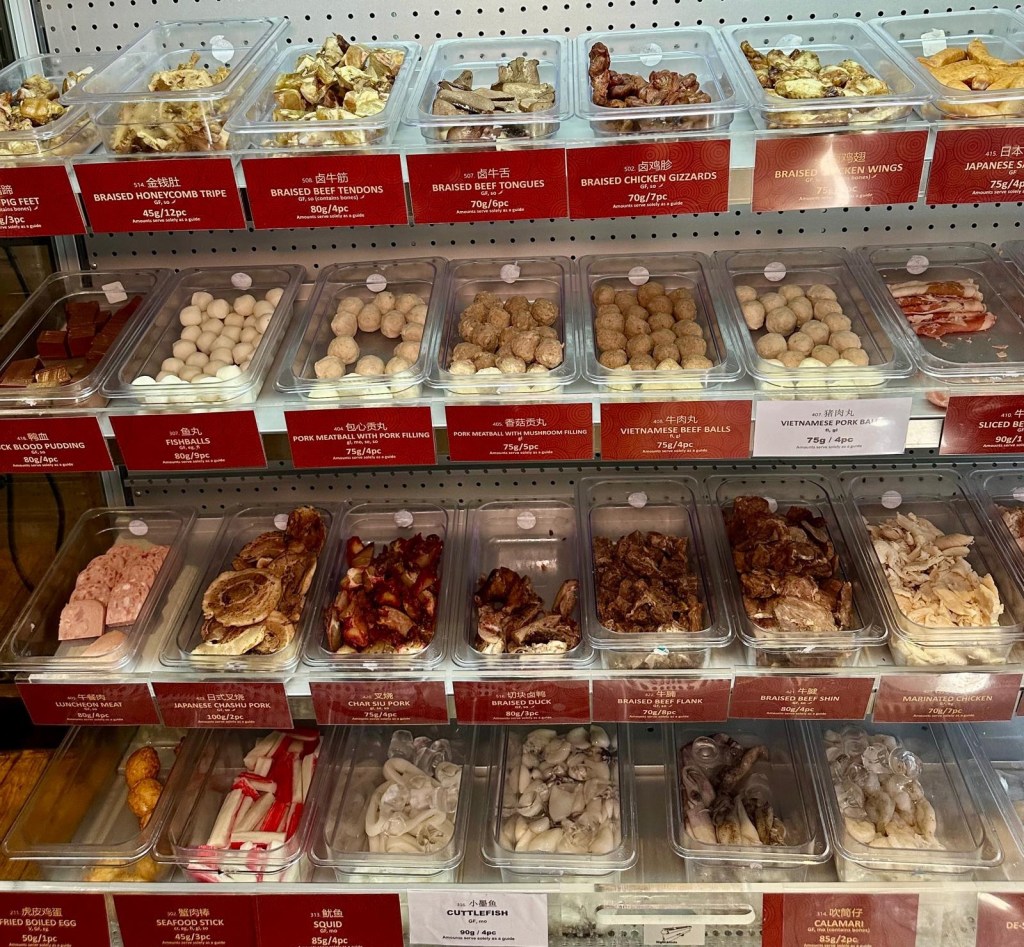 A row of refrigerated shelves stocked with fresh ingredients, including leafy greens, mushrooms, seafood, meats, and noodles, ready for selection.