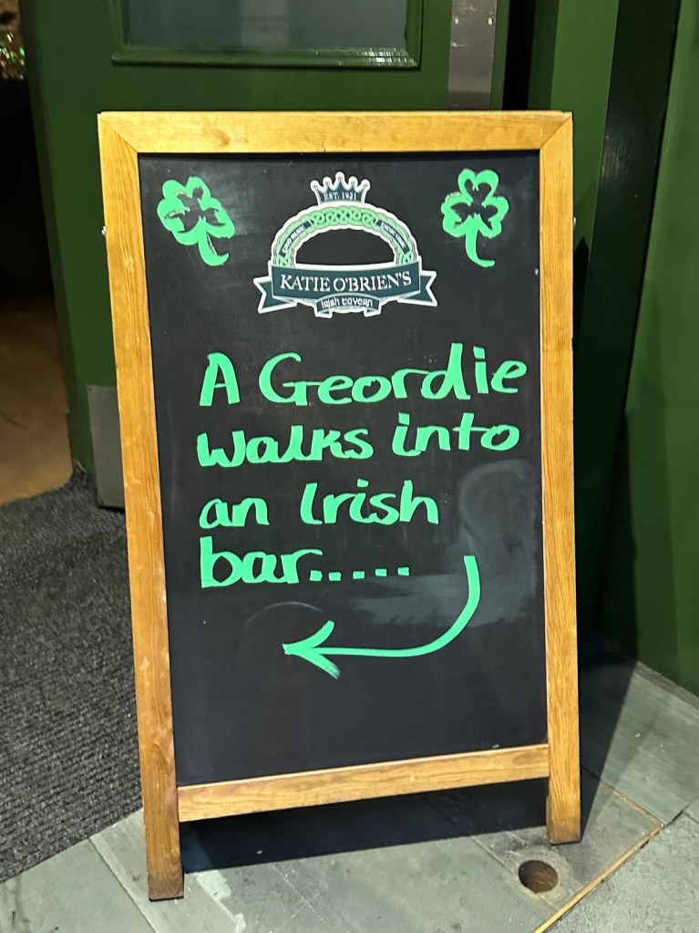 A sandwich board outside Katie O’Brien’s Irish bar in Newcastle, featuring a witty and humorous message welcoming guests.