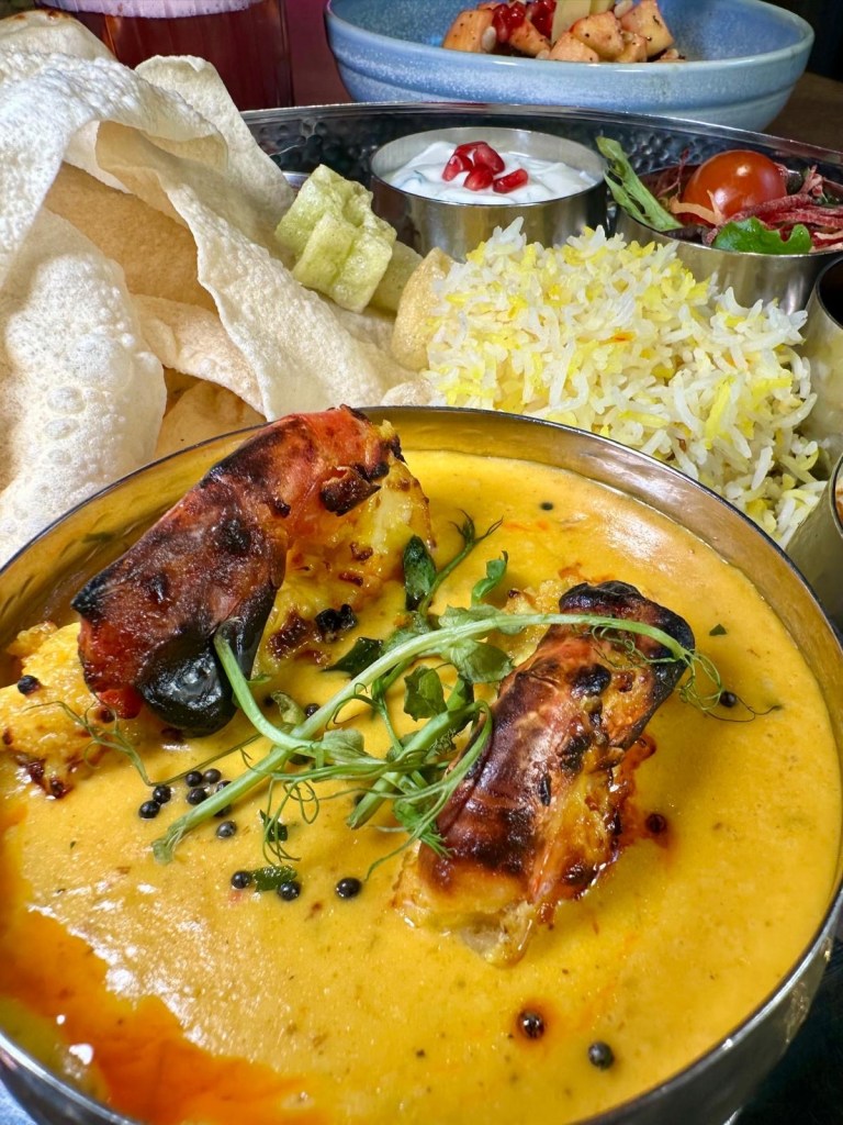 A vibrant thali platter featuring prawn moilee, saffron pulao, naan, curried black chickpeas, poppadoms, and a small gulab jamun.