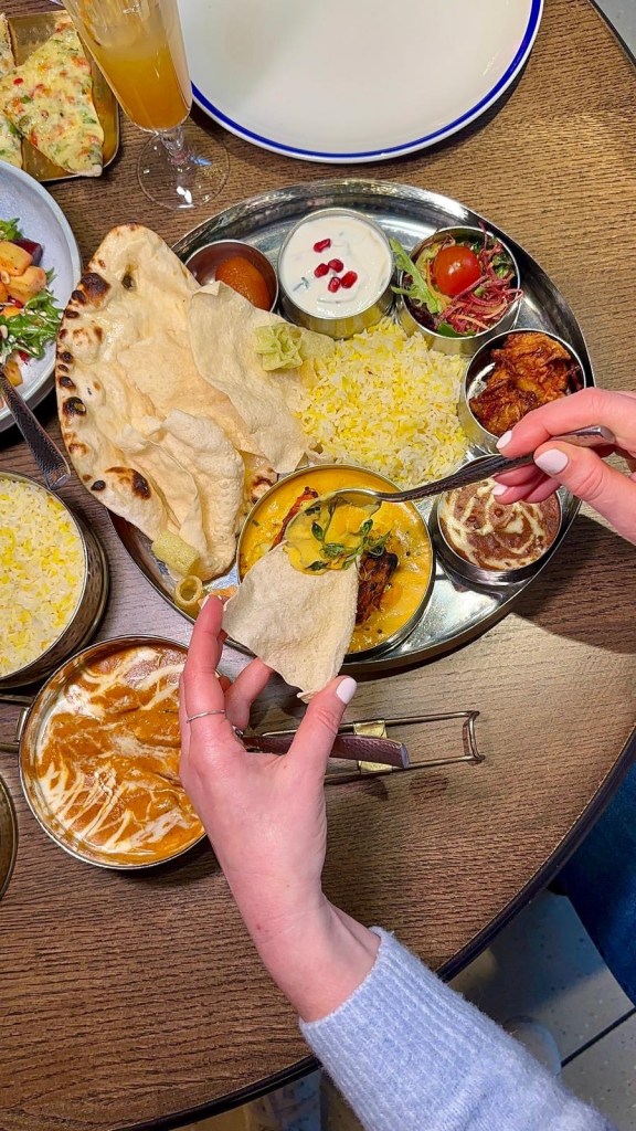 A vibrant thali platter featuring prawn moilee, saffron pulao, naan, curried black chickpeas, poppadoms, and a small gulab jamun.