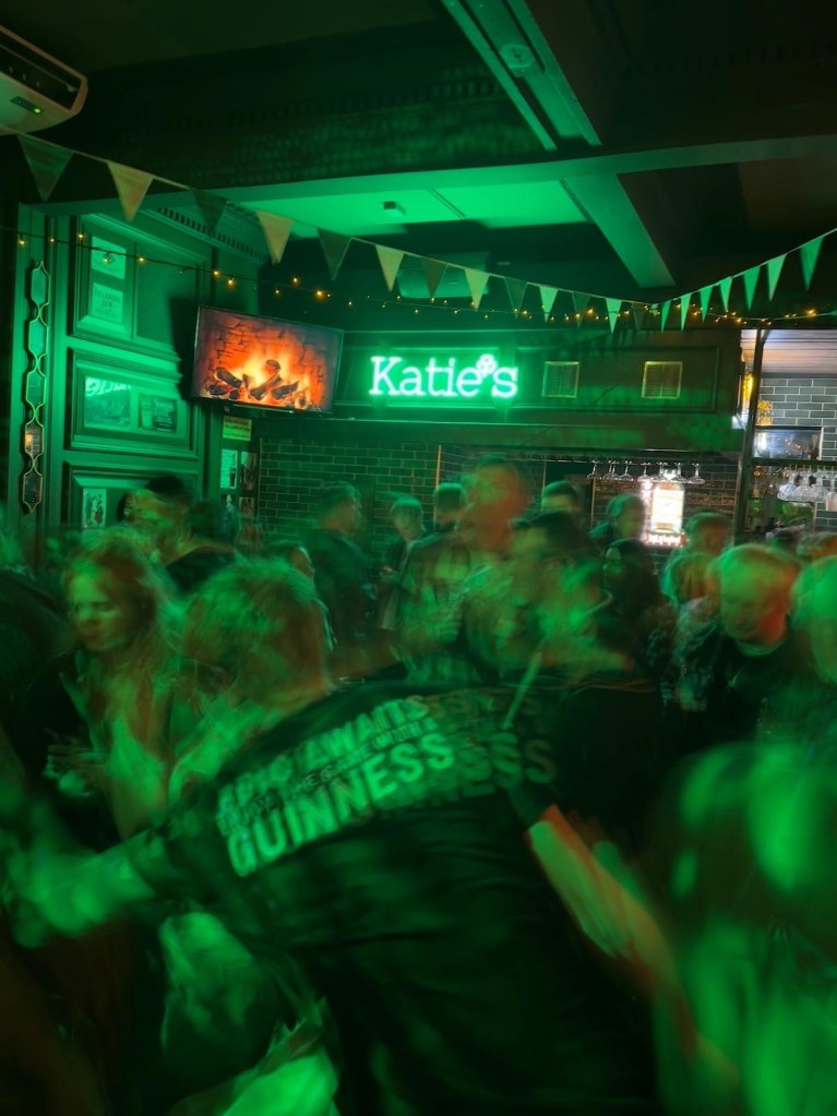 A spacious dance floor surrounded by rustic wooden tables and bar stools, filled with people enjoying drinks and music.