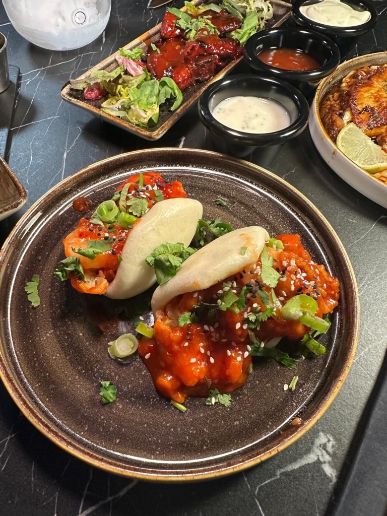 Two soft, fluffy bao buns filled with crispy dynamite prawns and drizzled with a flavorful sauce.