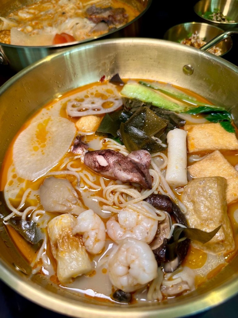 A steaming bowl of Malatang broth filled with fresh seafood, mushrooms, and Asian greens, garnished with coriander, crushed peanuts, and red chili.