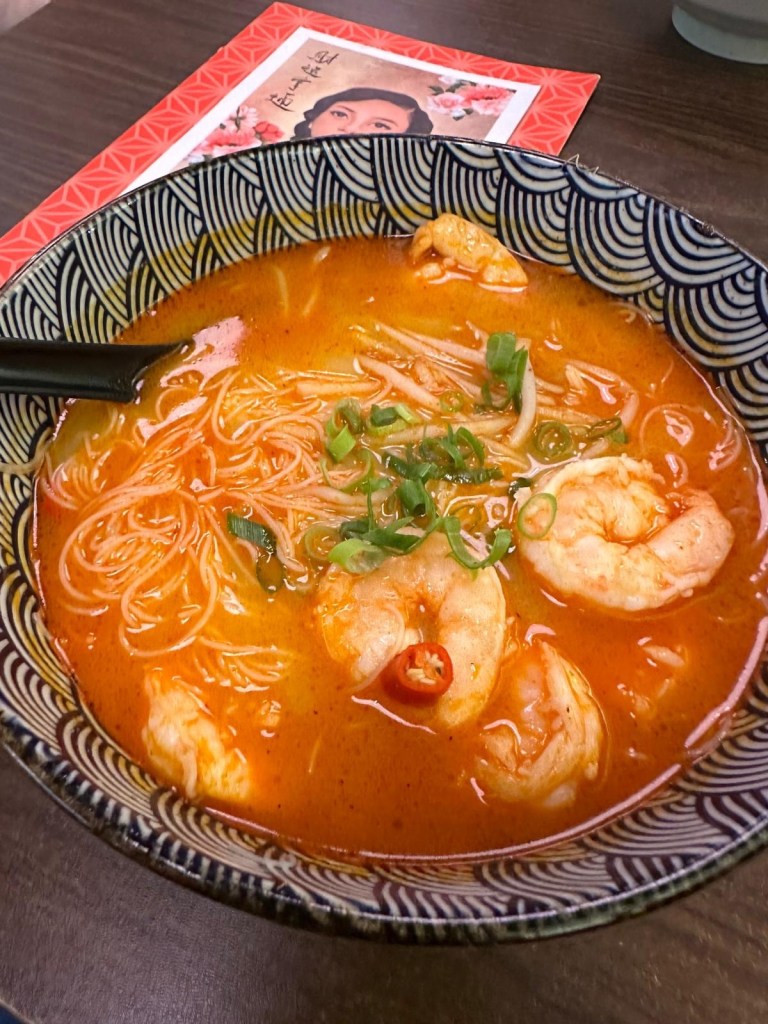 A bowl of prawn tom yum soup with a rich, aromatic broth, garnished with fresh herbs and served with a side of spicy oil.