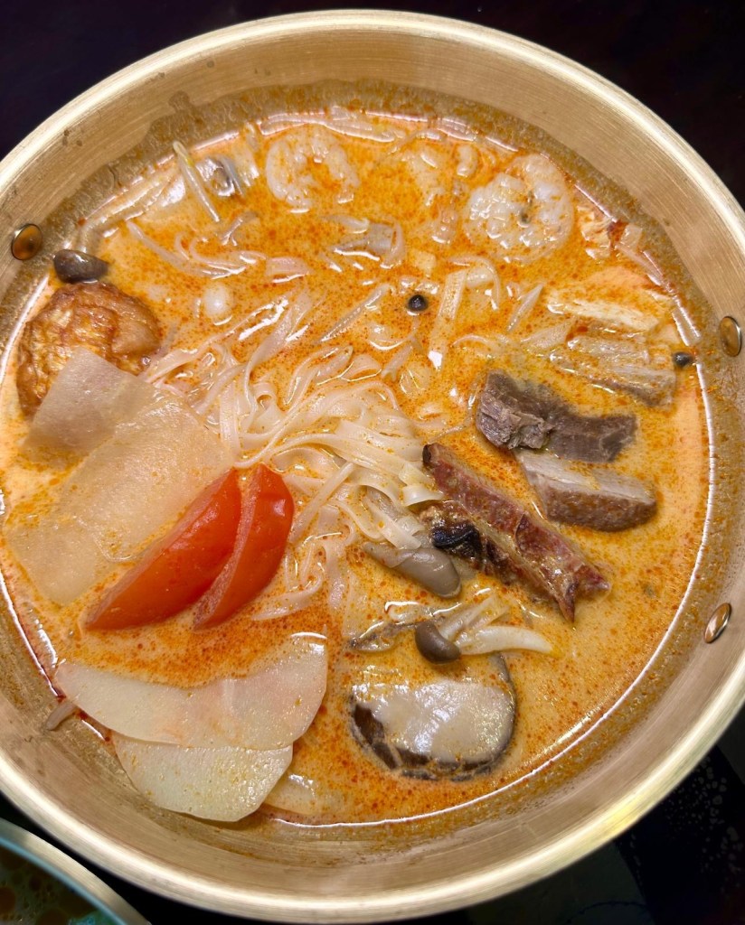 A vibrant bowl of Tom Yum soup with tender meats, mushrooms, and fresh vegetables, served with noodles and a side of chili oil.