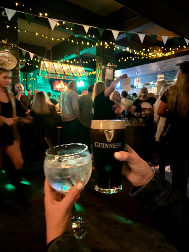 A spacious dance floor surrounded by rustic wooden tables and bar stools, filled with people enjoying drinks and music.