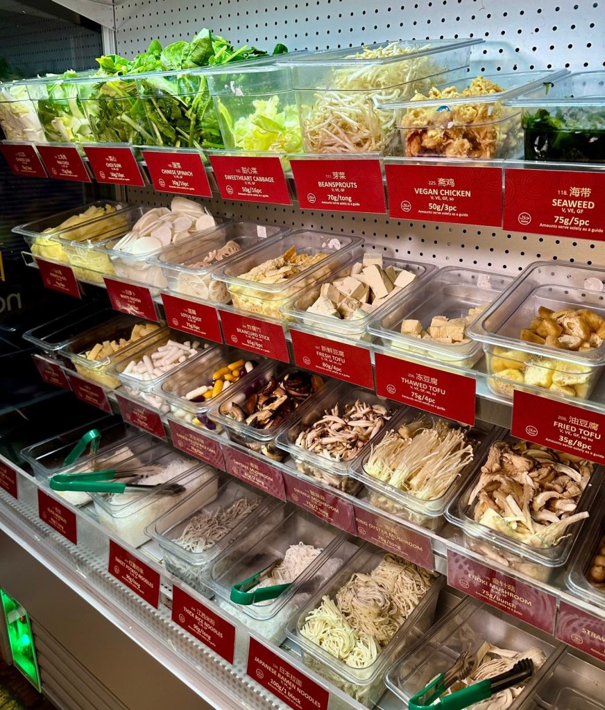 A row of refrigerated shelves stocked with fresh ingredients, including leafy greens, mushrooms, seafood, meats, and noodles, ready for selection.