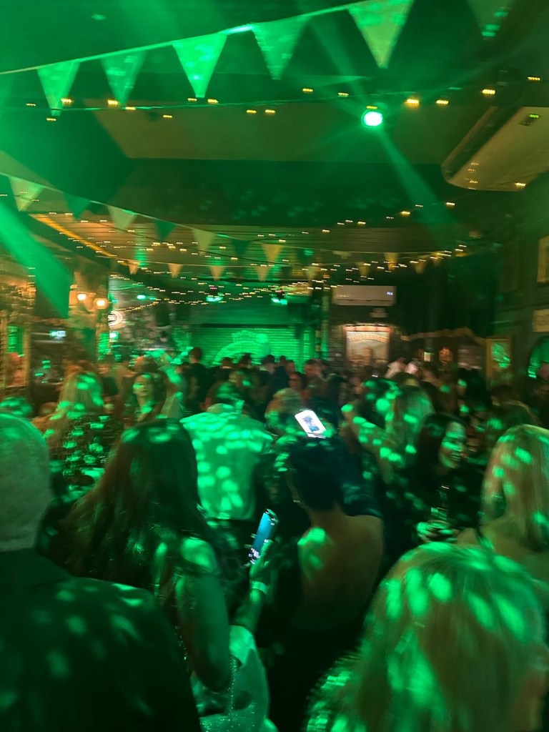 A spacious dance floor surrounded by rustic wooden tables and bar stools, filled with people enjoying drinks and music.