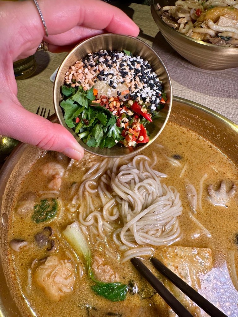 A creamy curry laksa broth with fresh seafood, leafy greens, and udon noodles, topped with a drizzle of chili oil and A peanut butter-infused dry noodle mix with thick udon noodles, mushrooms, and tender vegetables, tossed in a rich, nutty sauce.