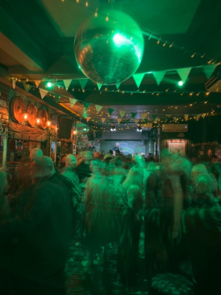 A spacious dance floor surrounded by rustic wooden tables and bar stools, filled with people enjoying drinks and music.