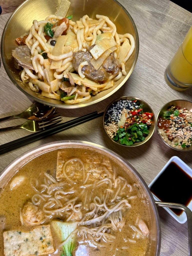 A creamy curry laksa broth with fresh seafood, leafy greens, and udon noodles, topped with a drizzle of chili oil and A peanut butter-infused dry noodle mix with thick udon noodles, mushrooms, and tender vegetables, tossed in a rich, nutty sauce.